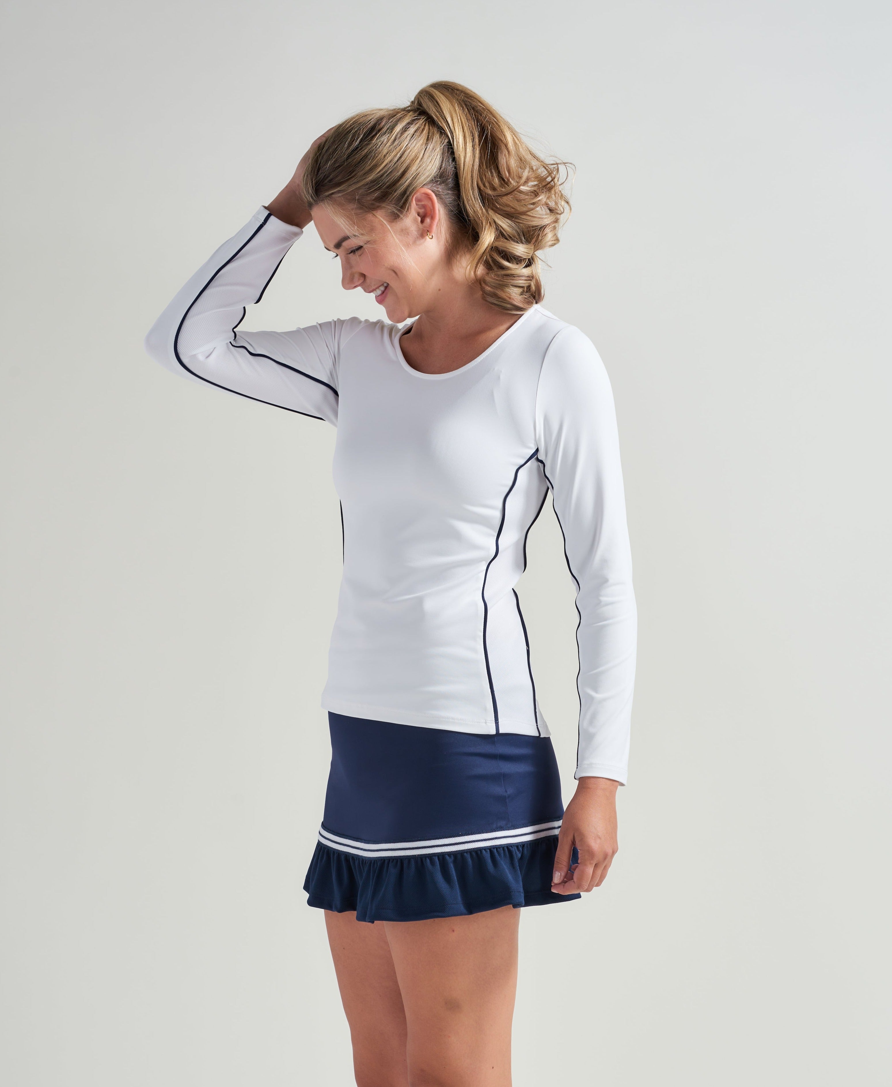 Woman tennis player wearing a long sleeve white tennis workout shirt with navy piping. Athlete is also wearing a navy tennis skort with navy and white knitted trim.