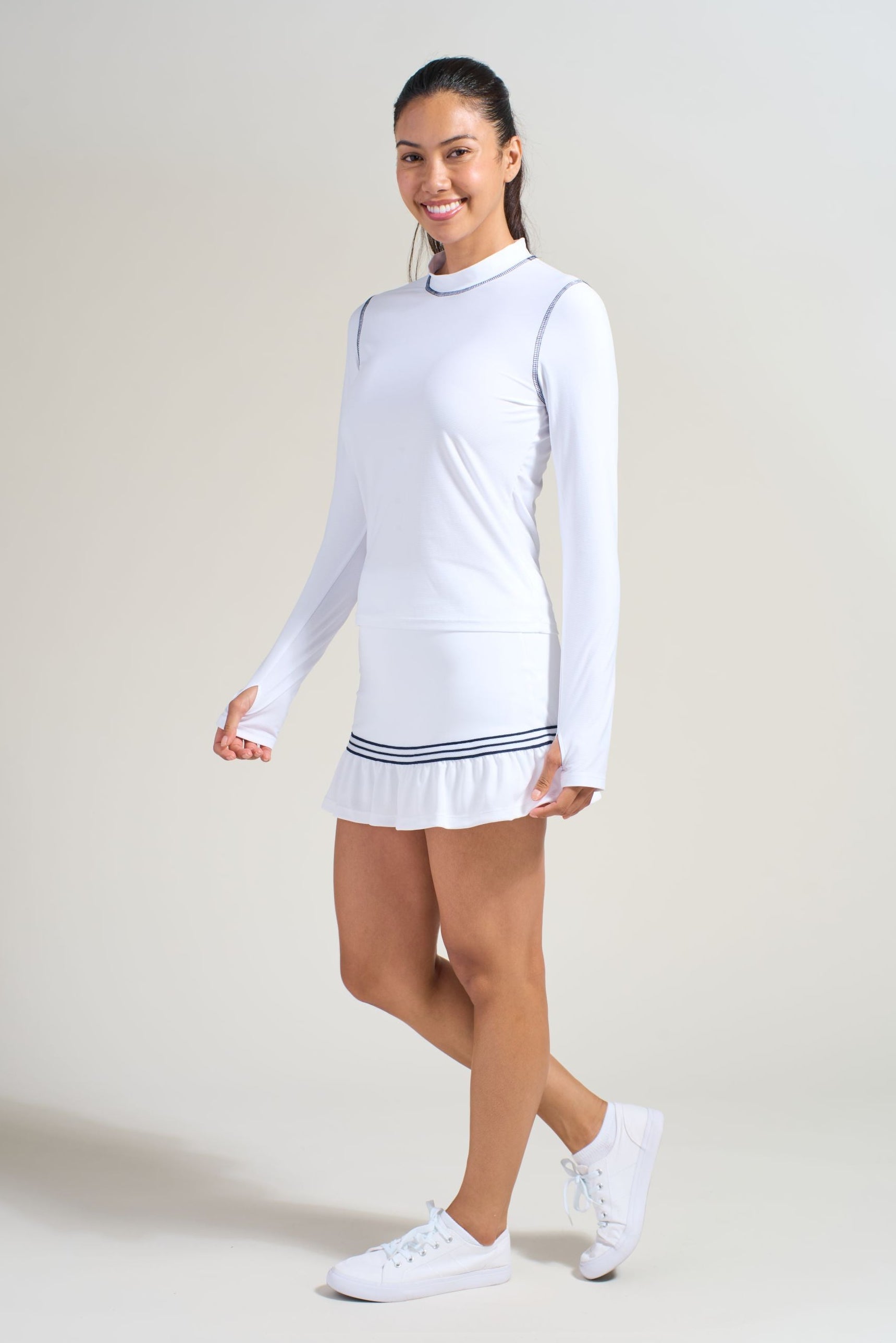 Women's athlete in all white tennis outfit with white long sleeve tennis shirt with navy stitching around armholes and collars. Also wearing a white tennis skort with lightweight mesh ruffles and navy trim.