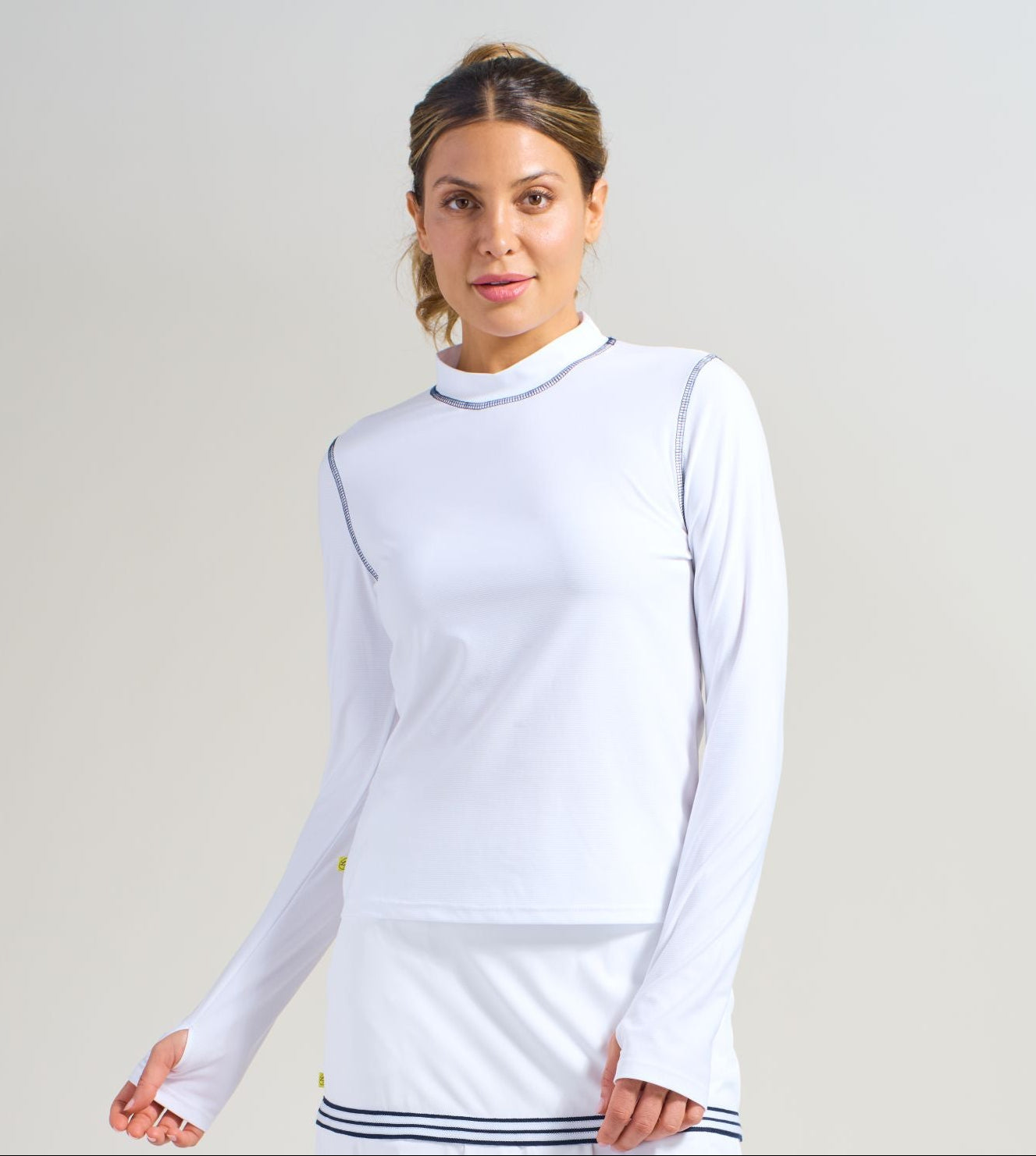 Women's athlete wearing long sleeve tennis shirt with navy trim around armhole and collar. Shirt also has thumbholes. Also wearing tennis skort with navy trim and lightweight ruffle mesh hem.
