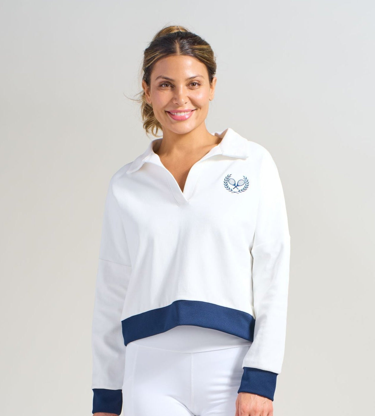 Women's tennis athlete wearing white cropped sweatshirt with navy ribbed cuffs and hem. Small tennis racquets embroidery on left side of chest. Also wearing matching white shorts with pockets.