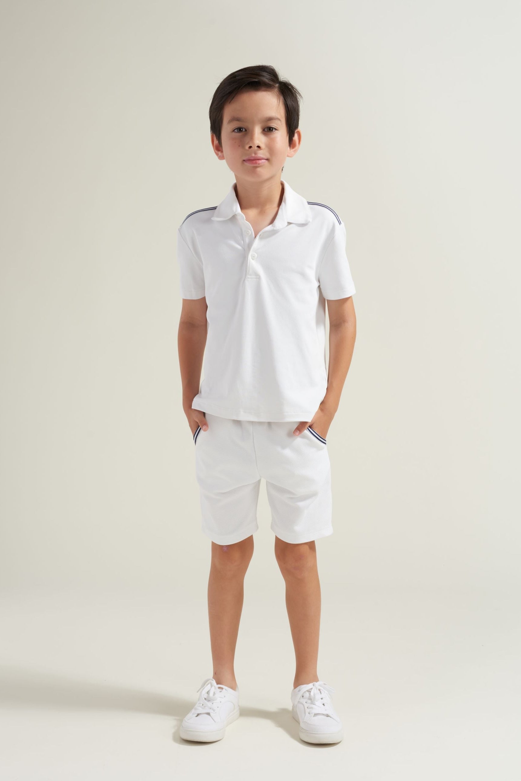 Youth tennis player wearing a white tennis polo shirt with navy and white trim on the shoulders. Youth tennis shorts with navy and white accents along pockets.