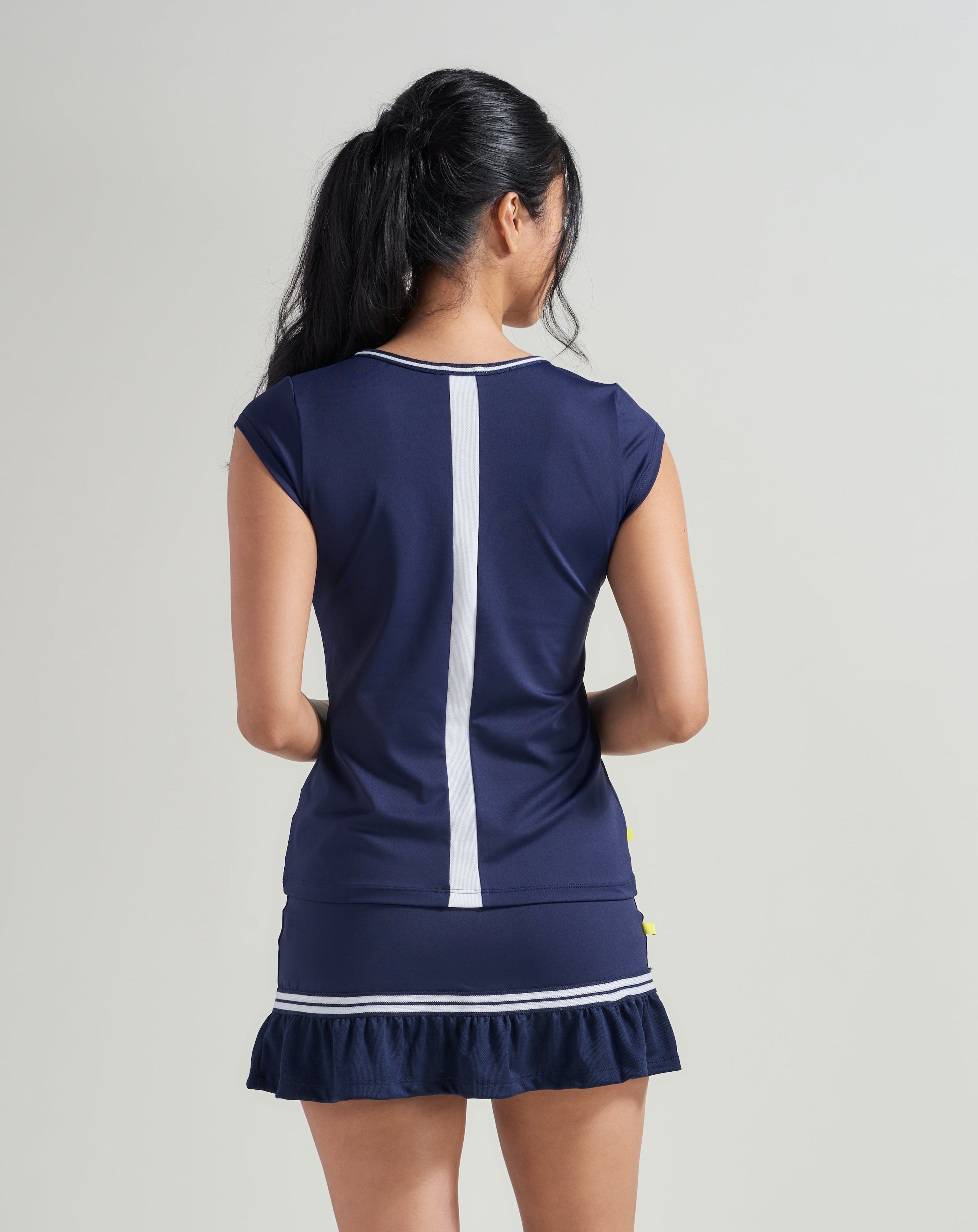 Back view of a women's tennis t-shirt with white mesh stripe down back and navy and white knitted trim along neckline. Woman is also wearing a navy women's skort with navy and white trim along navy mesh.