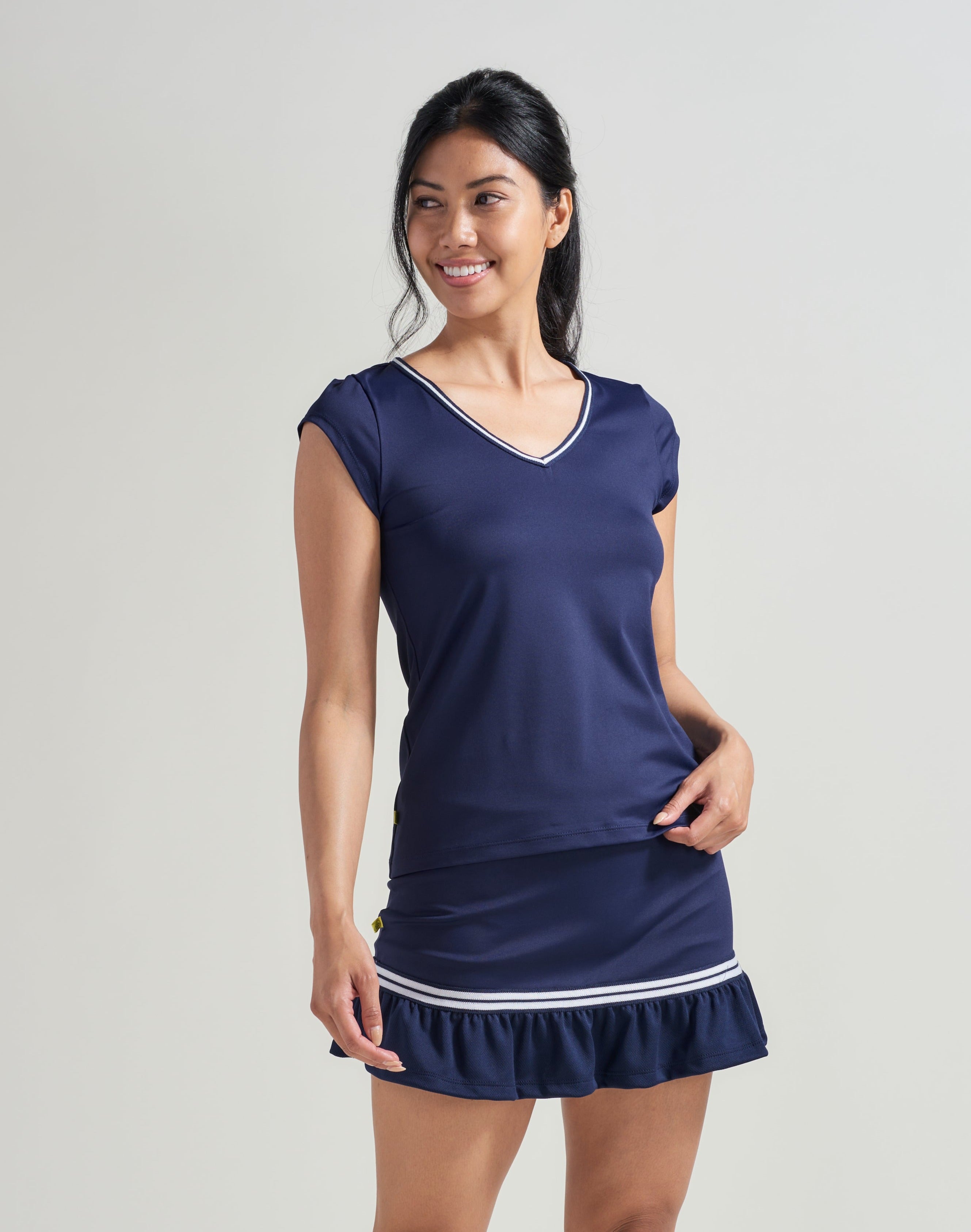 A female tennis player wearing a women's tennis V-neck t-shirt in navy with navy and white trim along the neckline. Woman is also wearing a navy tennis skort with navy and white knitted trim along navy mesh.