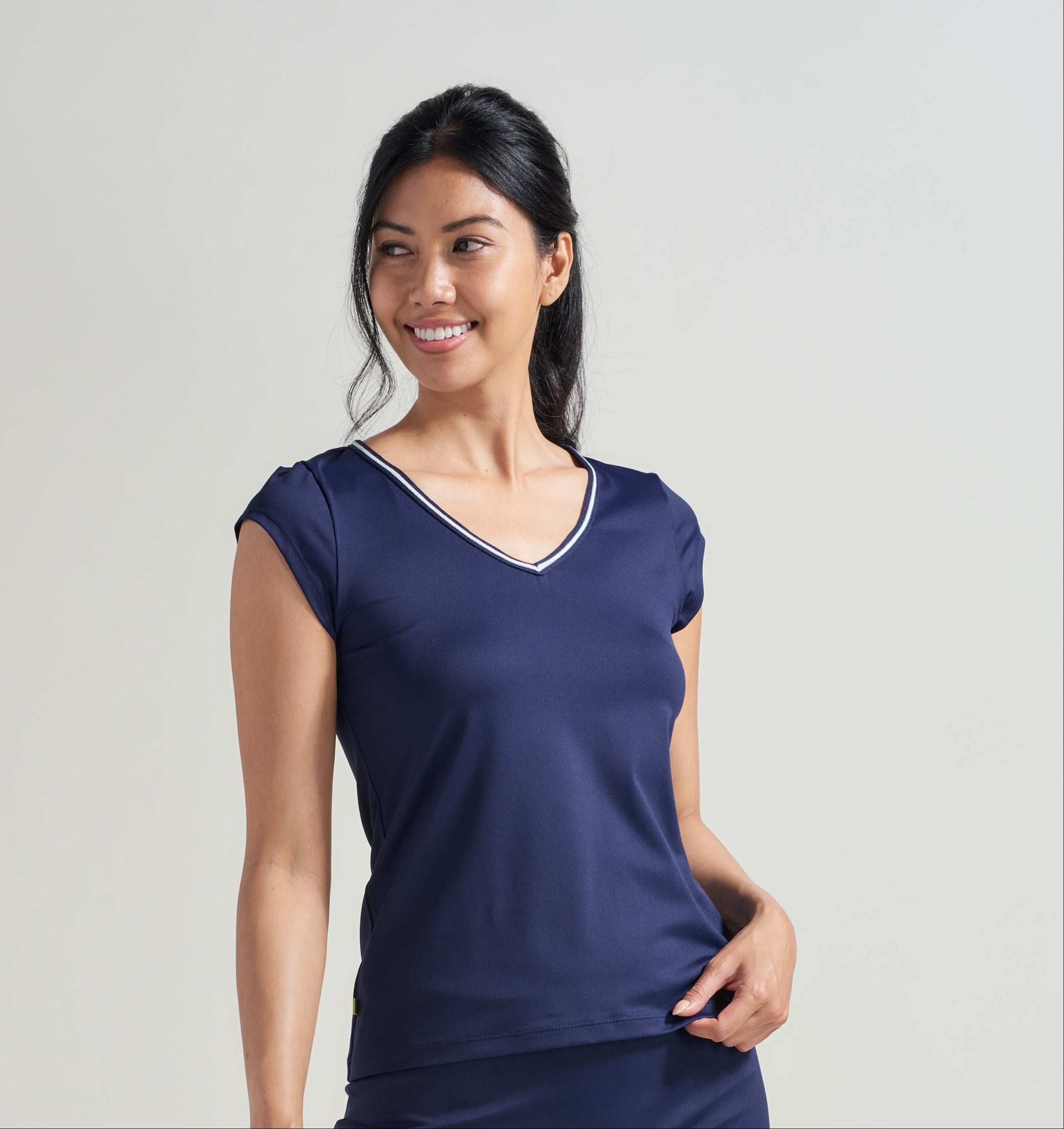 A female tennis player wearing a women's tennis V-neck t-shirt in navy with navy and white trim along the neckline. Woman is also wearing a navy tennis skort with navy and white knitted trim along navy mesh.