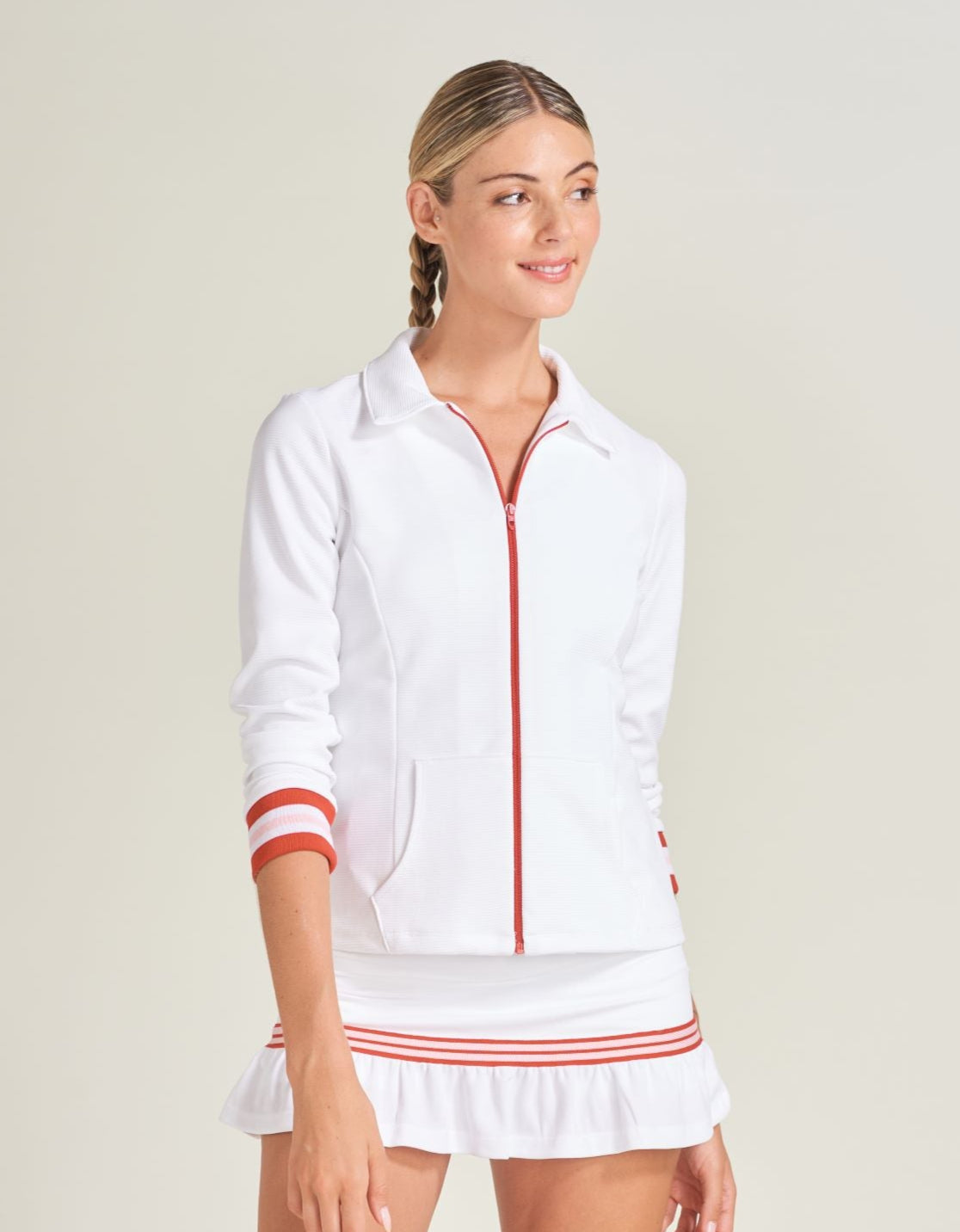 Woman tennis player wearing a white tennis warm up jacket with red and pink accents along ribbed cuffs. Woman is wearing a white tennis skort with red and pink knitted tape along white mesh.