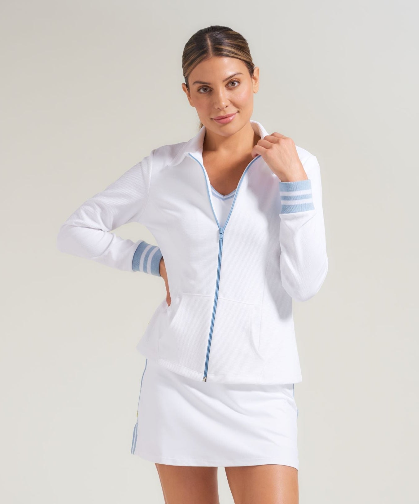 A woman tennis player wearing a white tennis workout jacket with blue zipper and blue and white ribbed cuffs on jacket. Woman is also wearing a white tennis skort with blue knitted tape along side seam and side slit.