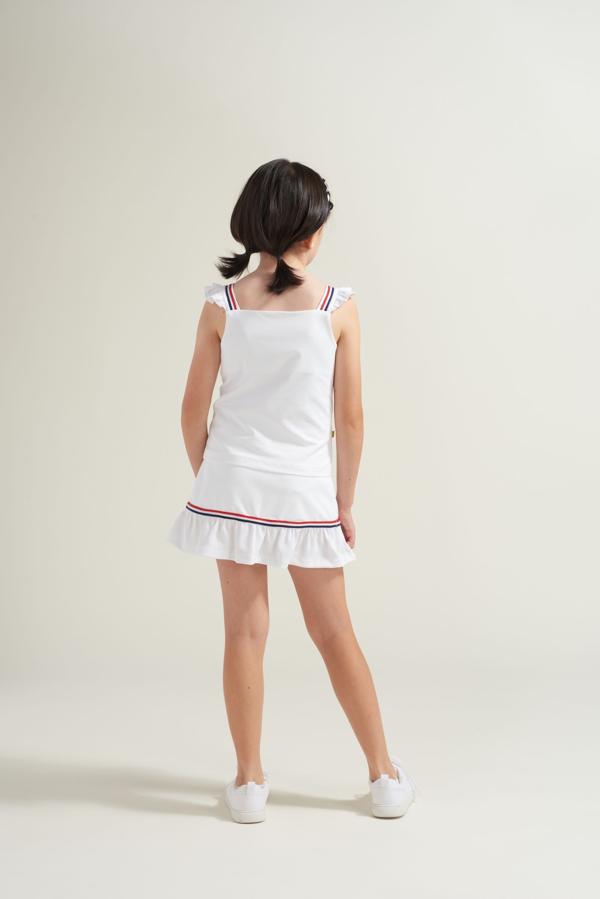 Back view of all white kids tennis shirt and tennis skort with red and blue trim.
