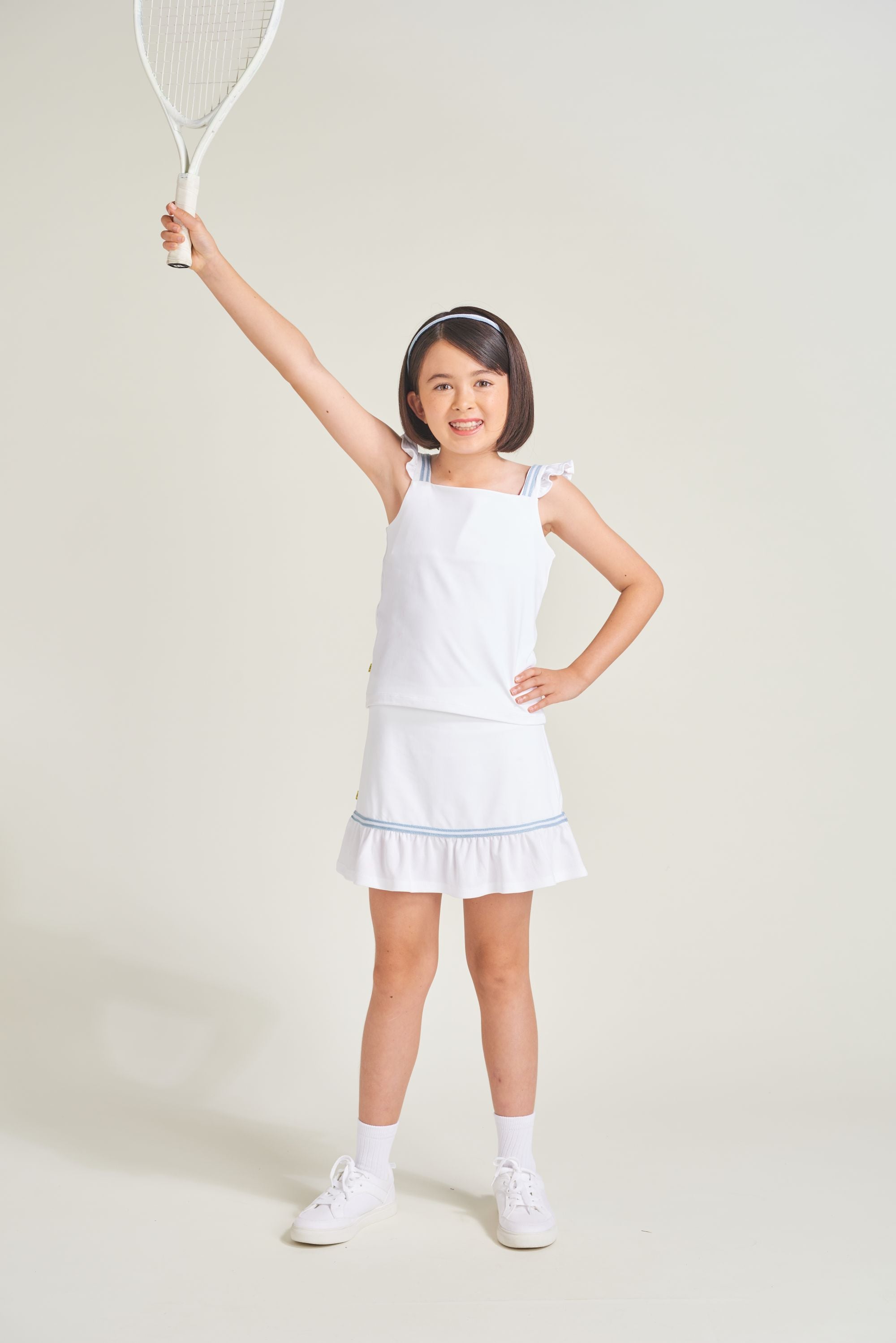 Girls youth tennis athlete is wearing a white tennis skort with shirring and blue and white trim. Girl is also wearing matching tennis tank top.