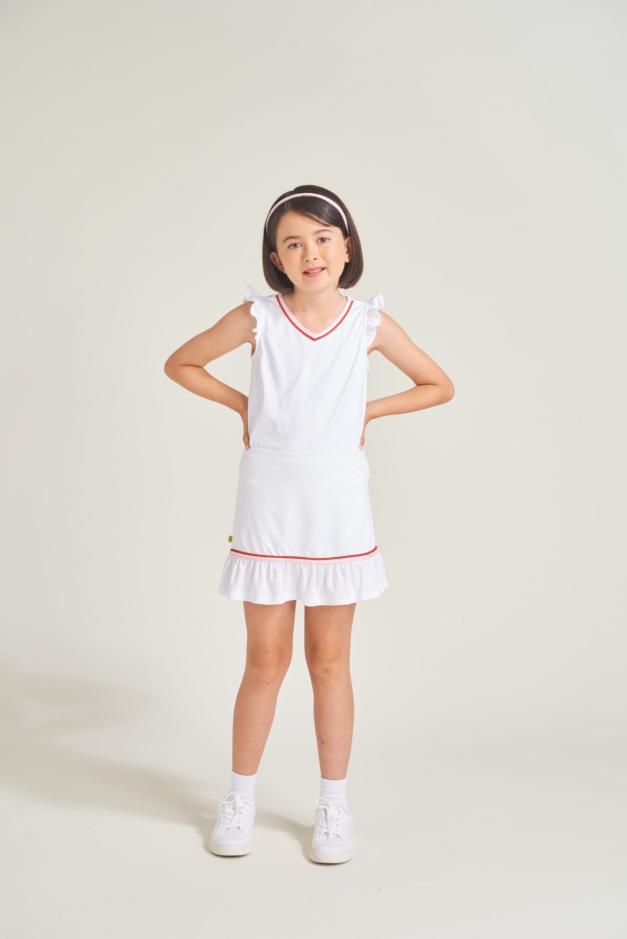 Youth girls tennis athlete is wearing a matching white tennis set. Girl is wearing a white skort with shirring and a knitted red and pink accent with a matching white tennis t-shirt with red and pink trim on the neckline.
