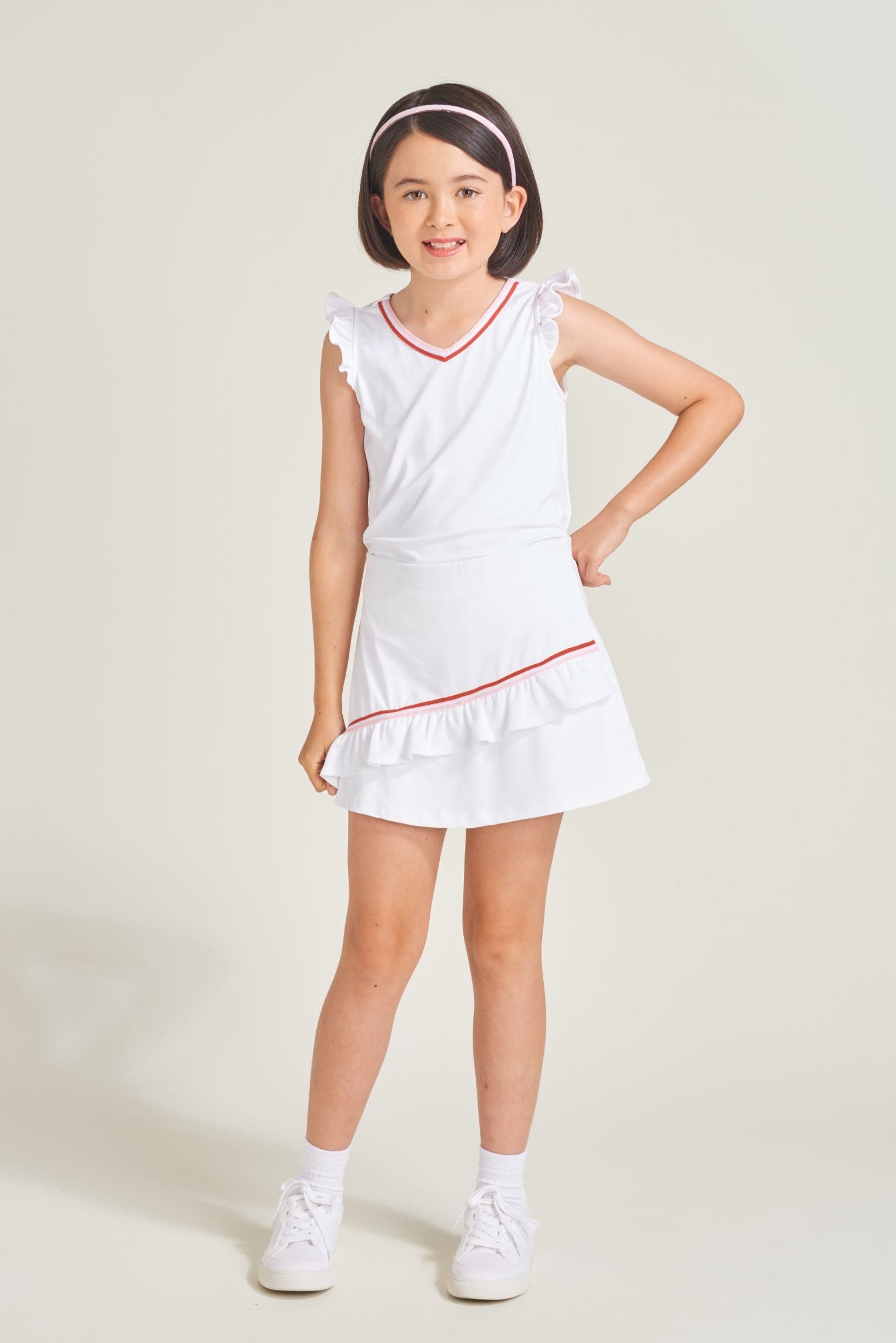 Youth tennis girl wearing matching white tennis set. Girl is wearing a white tennis skort with an asymmetrical ruffle with red and pink trim.  Youth girl is wearing matching sleeveless ruffle tennis t-shirt for girls.