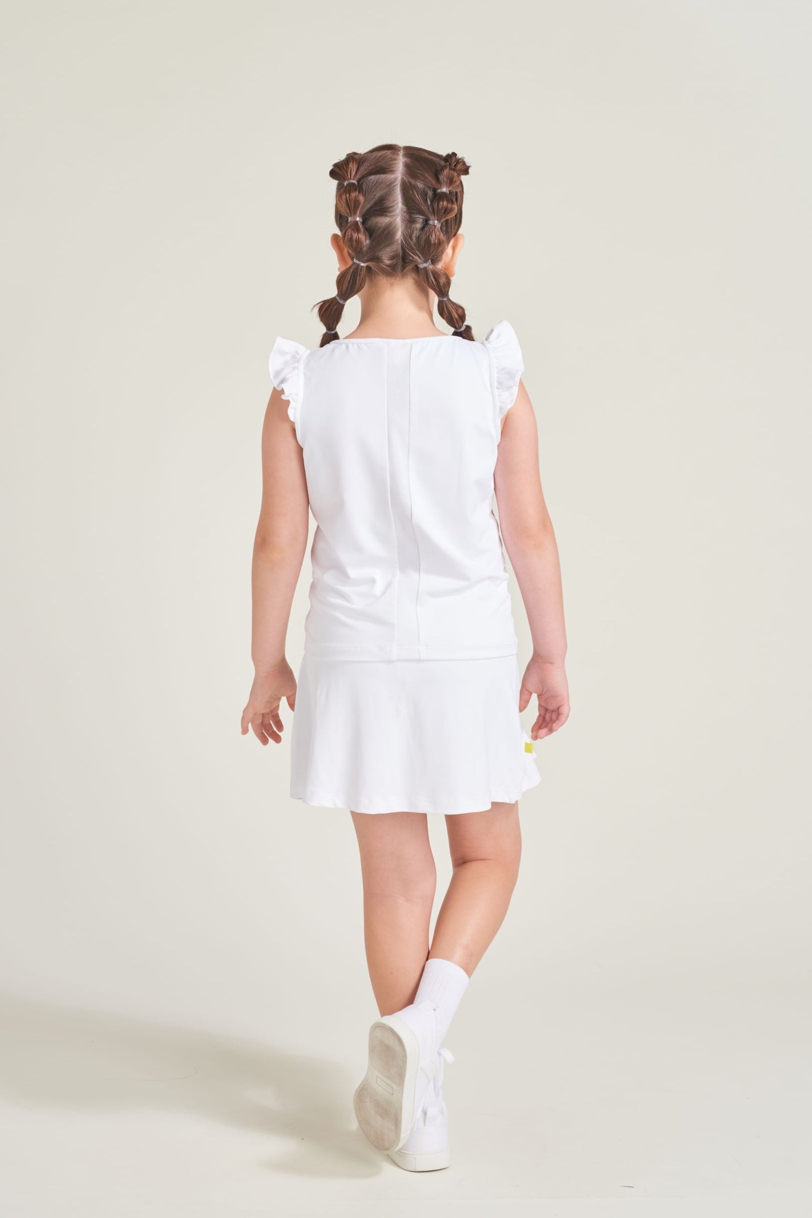 Back view of youth girls' white tennis tank top with white mesh back strip for breathability and ruffled sleeves. Girl is also wearing matching white tennis skort.