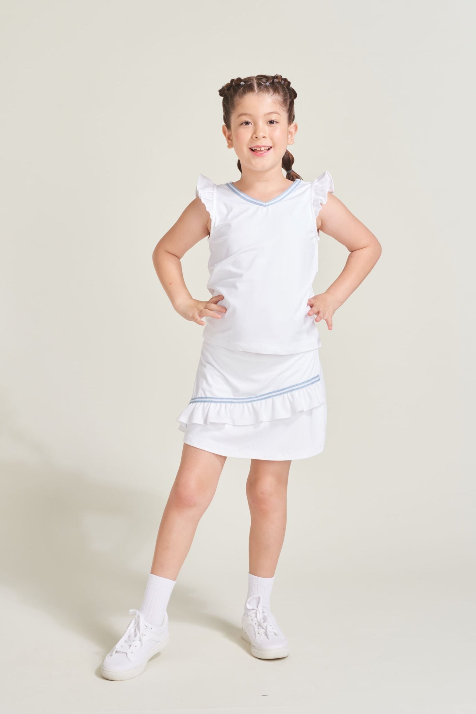 Youth girls tennis player wearing a white matching tennis tank top with blue and white trim along neckline and ruffled sleeves. Girl is wearing matching white tennis skort with asymmetrical ruffle and blue and white trim.
