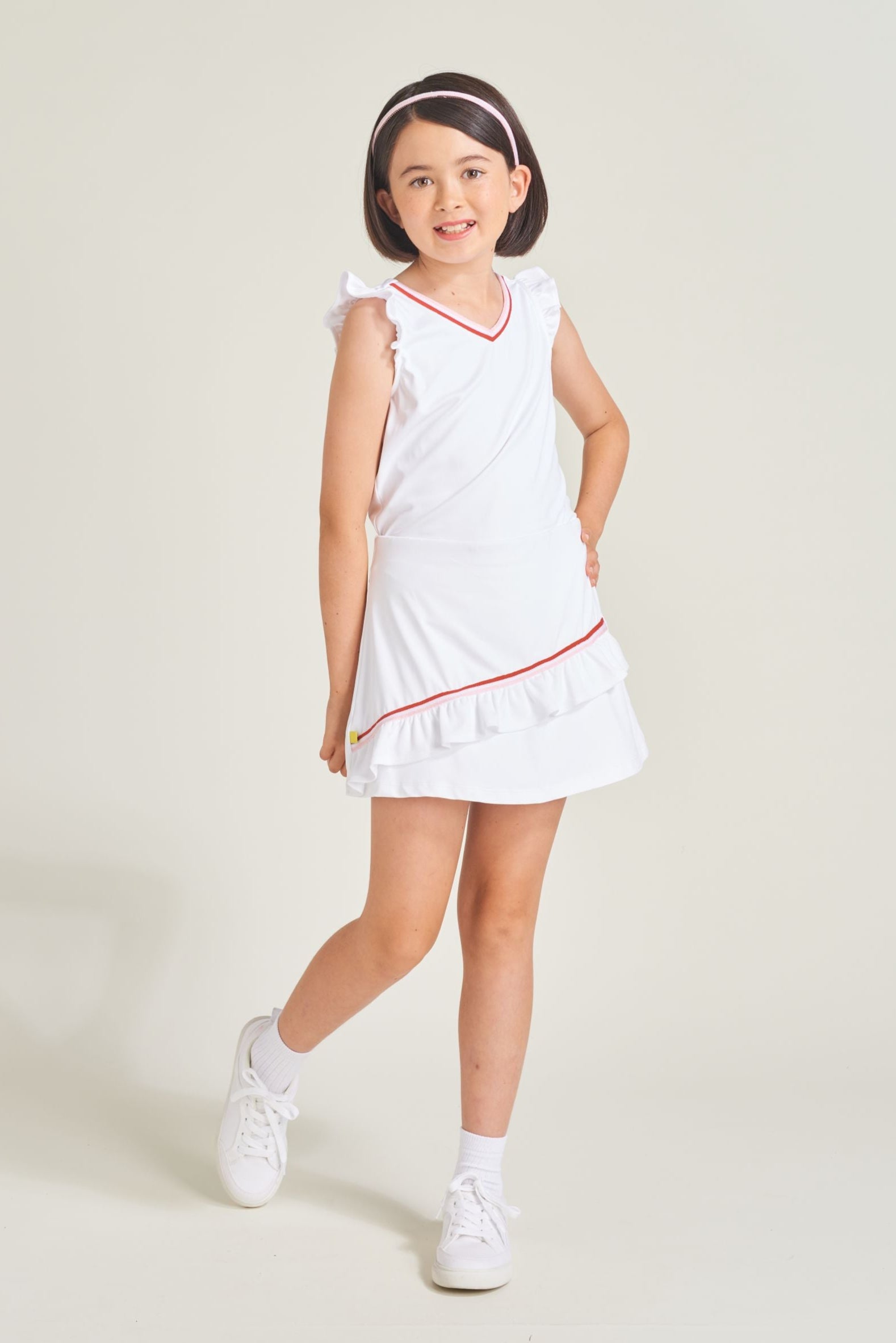 Youth girls tennis athlete is wearing a white tennis tank top with a match white tennis skort. The set has red and pink trim along the neckline and skort ruffle.