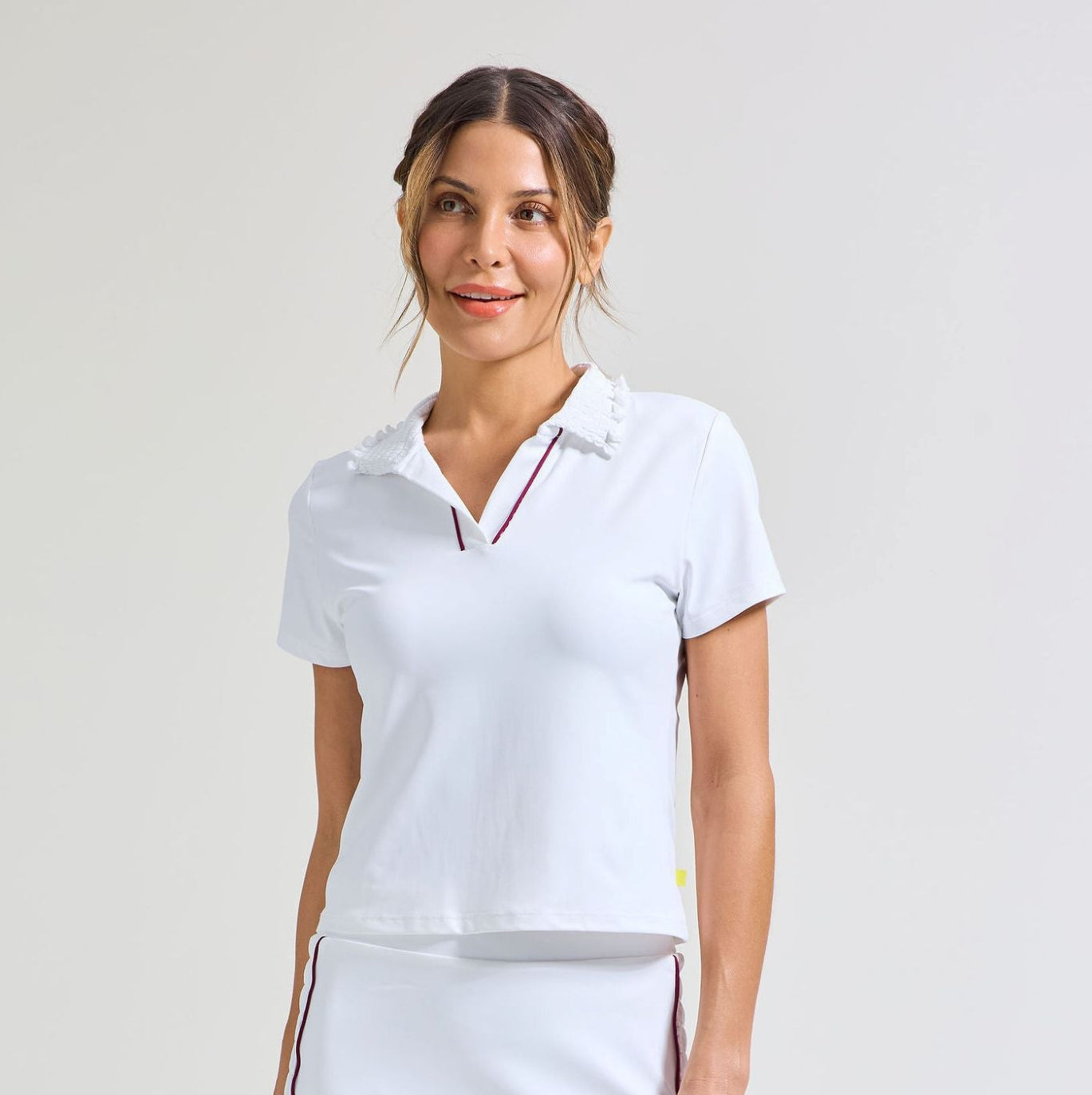 Women's tennis players in white polo shirt with smocked collar and burgundy piping along placket. Also wearing a matching white tennis skort with burgundy trim along side seams and smocking panel.