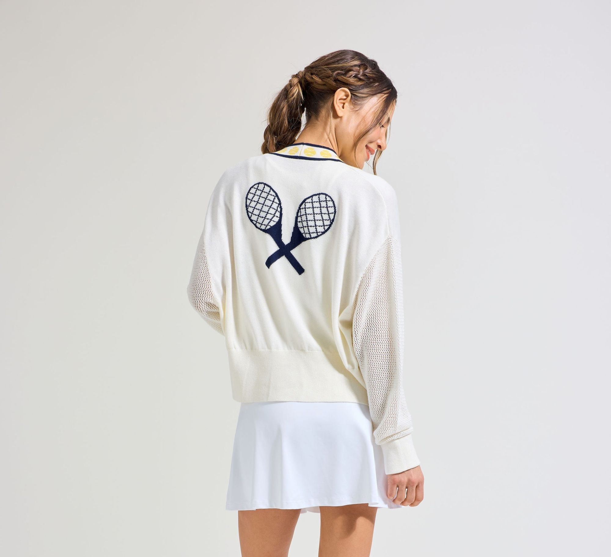 Back view of women's tennis player in white tennis cardigan with navy tennis embroidery on the back. Wearing matching white tennis skort.