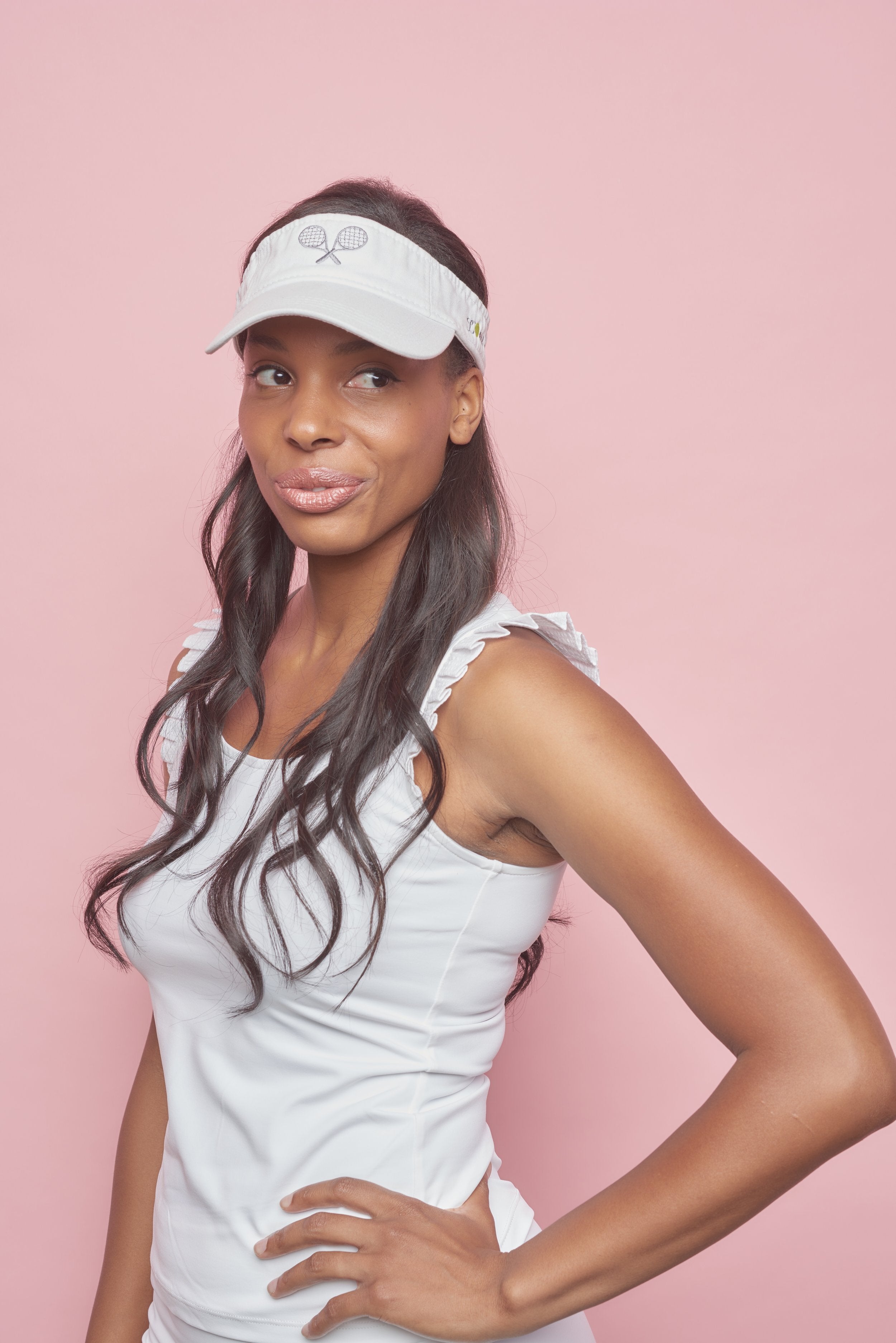 Woman tennis player in white women's tennis visor with grey embroidered tennis racquets. Woman is also wearing women's tennis t-shirt with pleated sleeves.