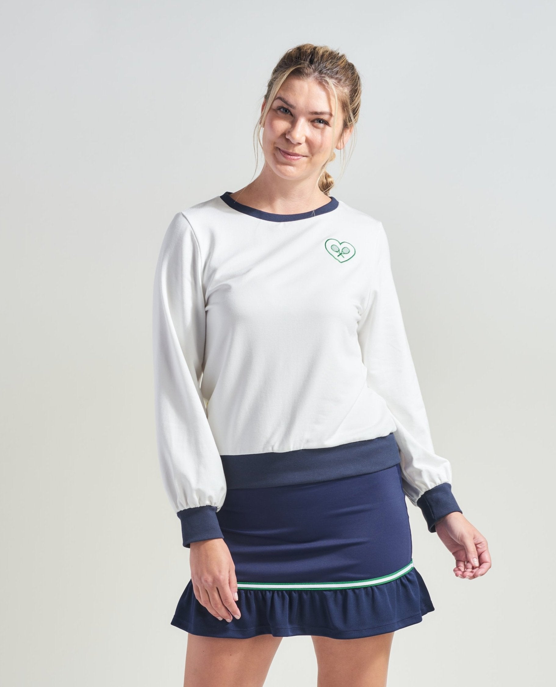 Woman tennis player is wearing lifestyle white tennis sweatshirt with green dainty accents and navy accents. Woman is also wearing  navy tennis skort for women.