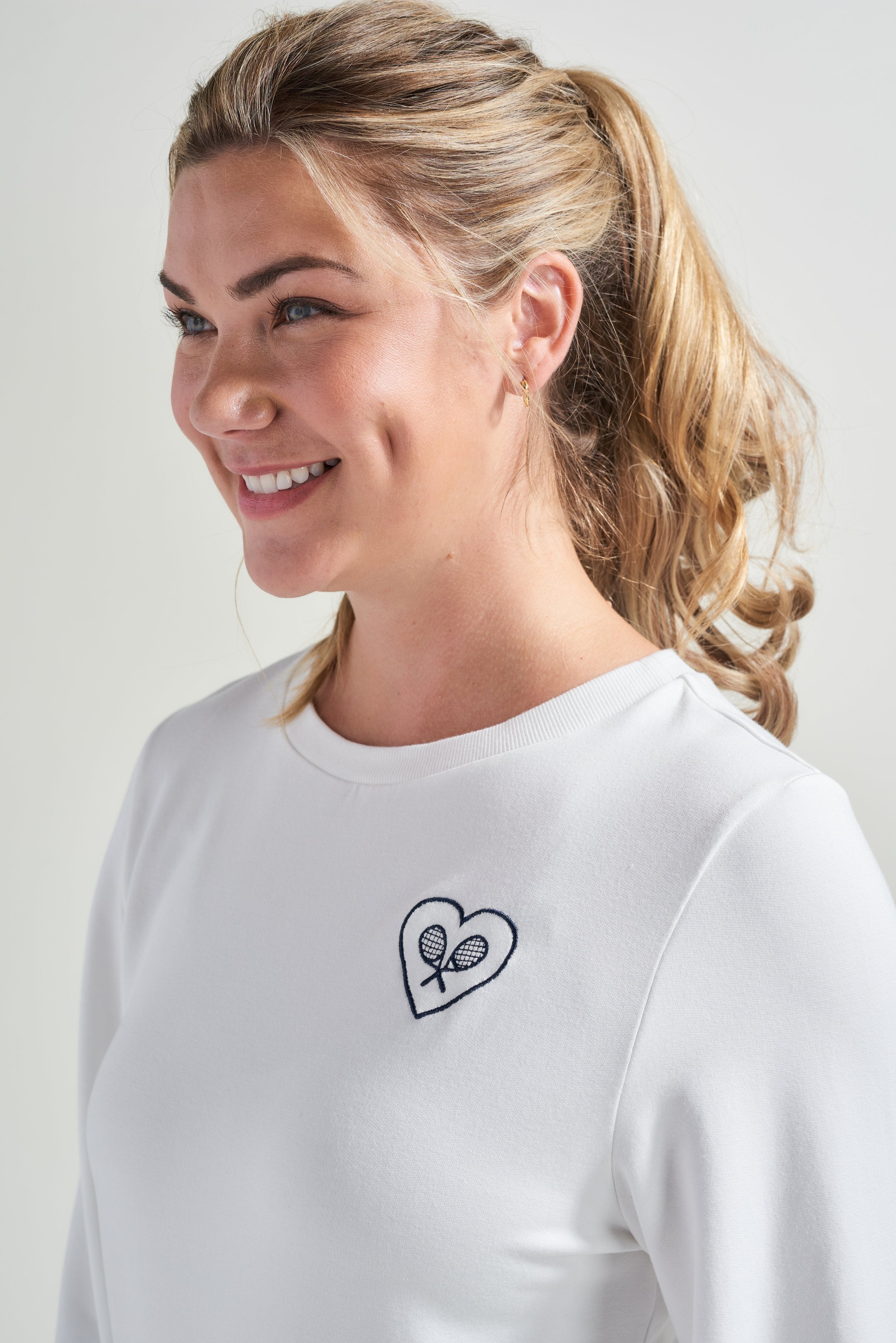 Detail picture of woman tennis player wearing a white sweatshirt with navy tennis embroidery accent.