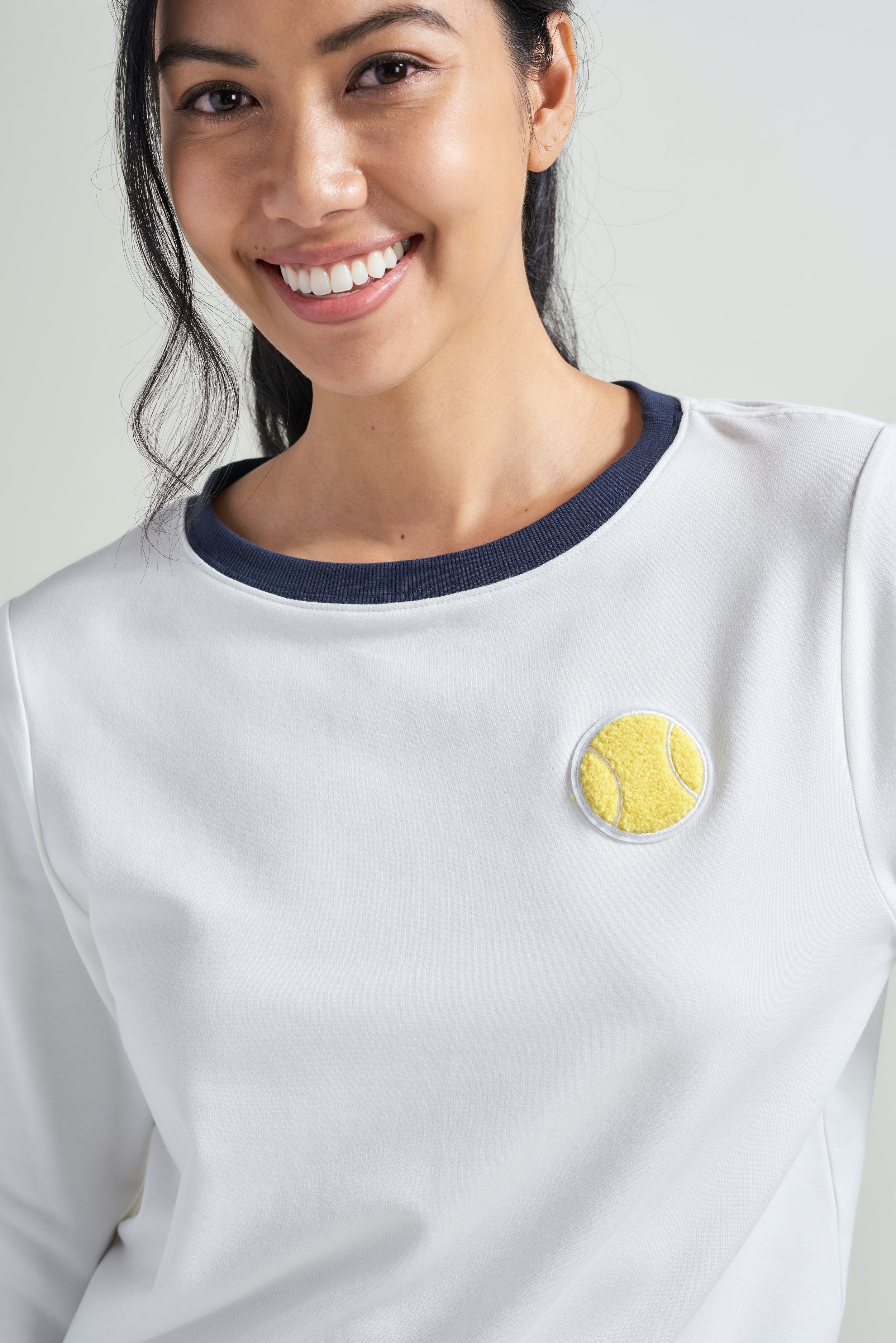Detail picture of female tennis player in women's white tennis sweatshirt. Tennis sweatshirt has navy ribbed neckline and tennis embroidered patch on chest.