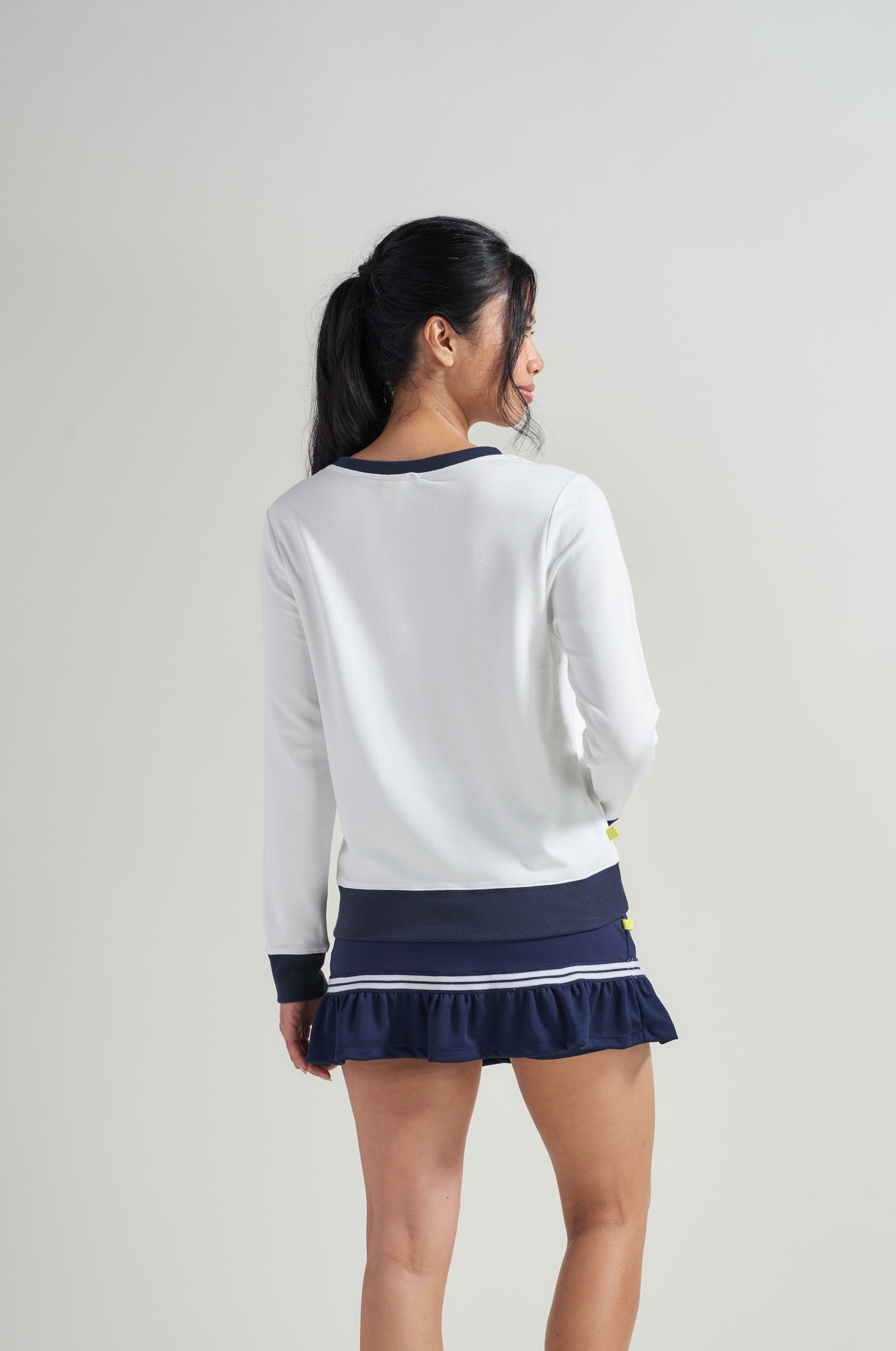 Back view of woman tennis athlete in white tennis sweatshirt and navy tennis skort.