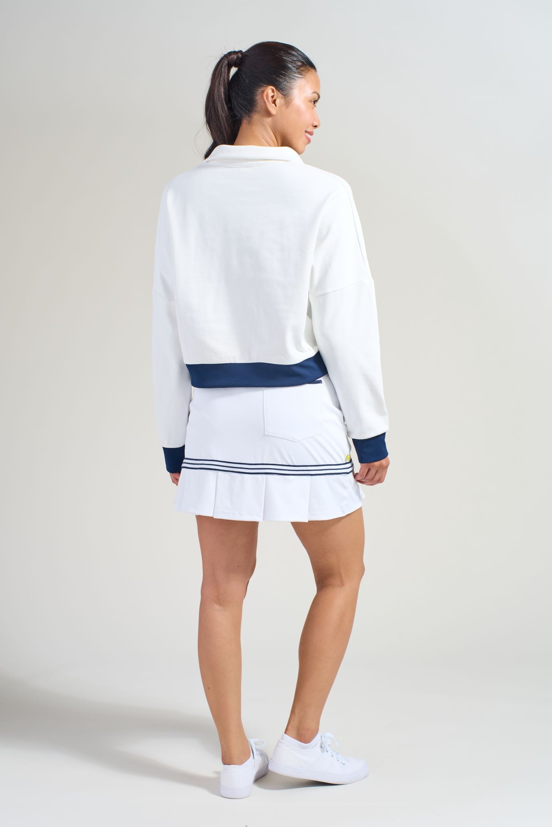 Back view of women's golf athlete wearing white and navy cropped sweatshirts with matching lightweight white golf skort with pleats around hem. 