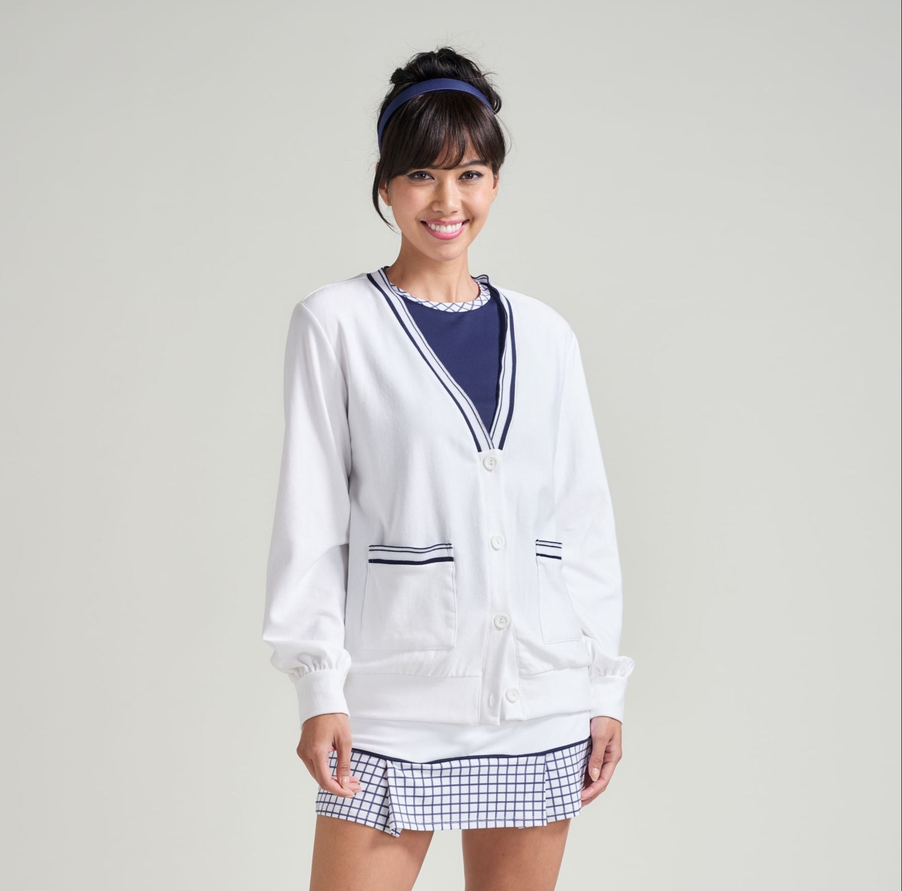 Woman wearing a white cardigan, navy shirt, plaid skirt, and white sneakers on a plain background